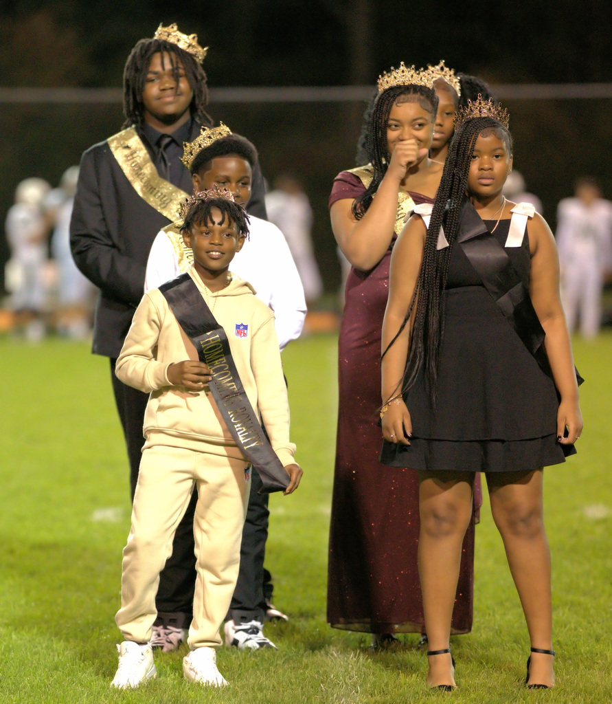 Members of the homecoming court stand together on the football field wearing formal attire, crowns, and ceremonial sashes.