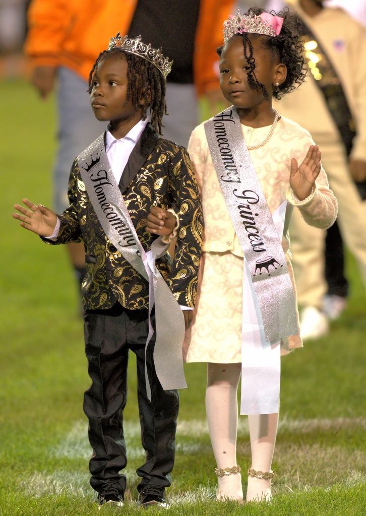 Two young children wearing crowns and silver homecoming prince and princess sashes stand together on the football field, holding hands and waving.