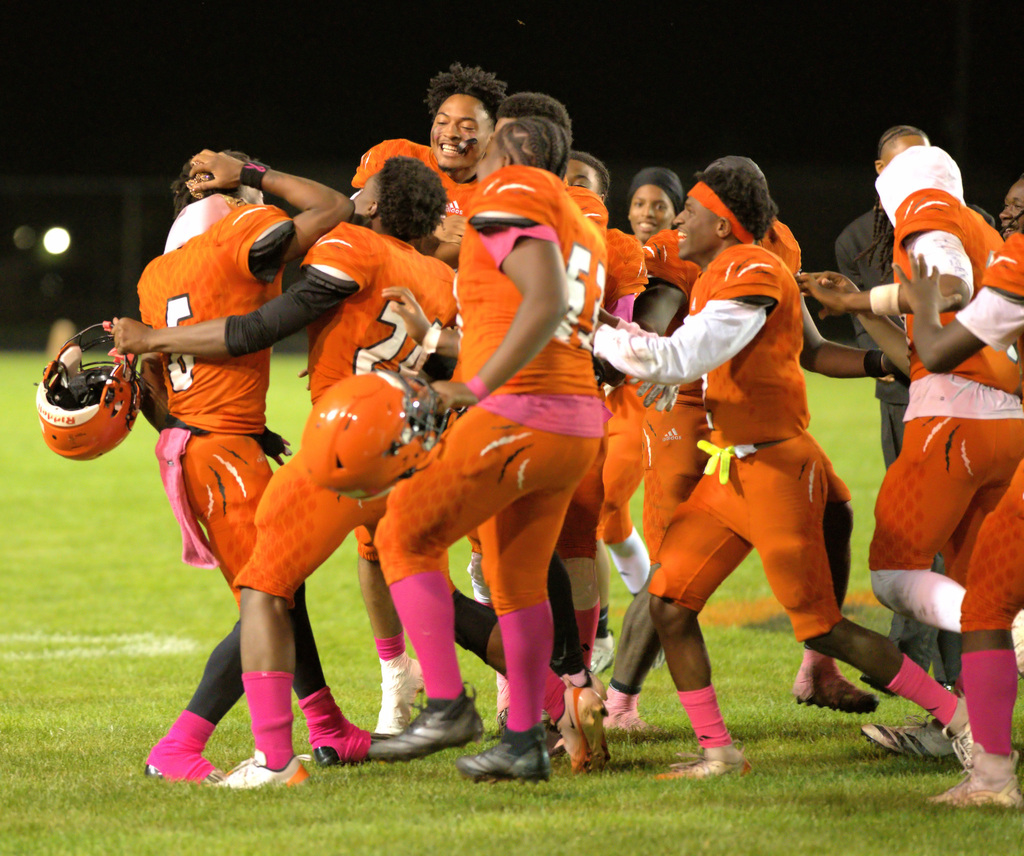 Benton Harbor Tigers football players in orange uniforms and pink accessories gather in a circle, smiling and celebrating the homecoming king on the field.