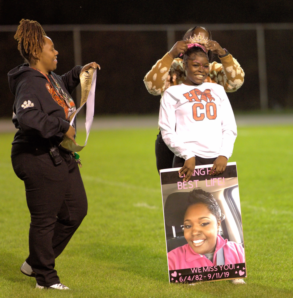 A smiling homecoming queen receives her crown during a ceremony on the football field while holding a memorial poster honoring a loved one.