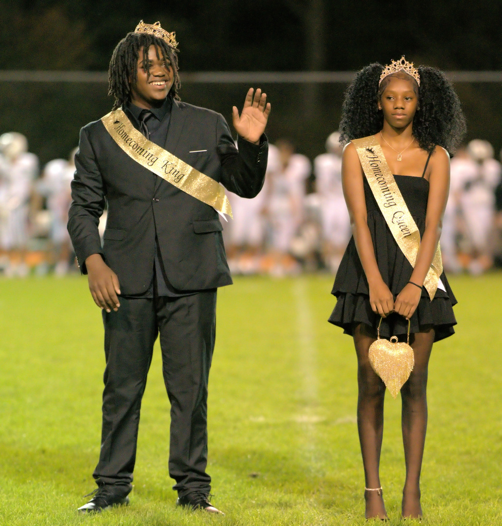 The middle school homecoming king and queen stand together on the football field wearing crowns and gold sashes. The king waves while the queen holds a gold heart-shaped purse.