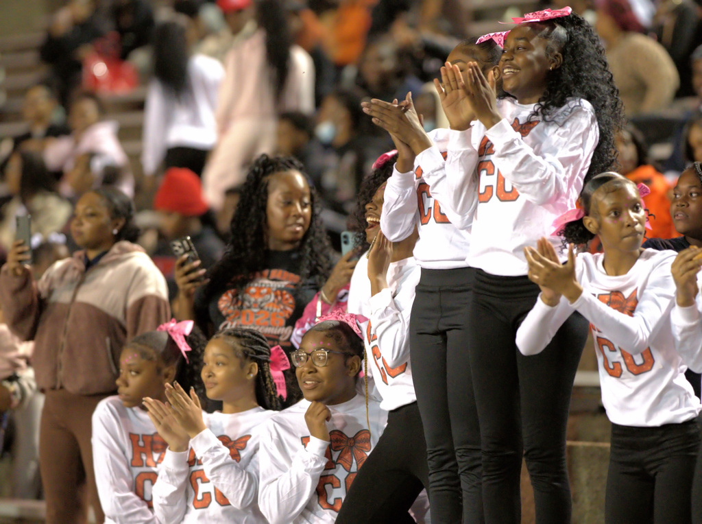 Members of the homecoming cheer squad, dressed in white shirts with orange lettering and pink bows, clap and cheer from the bleachers.