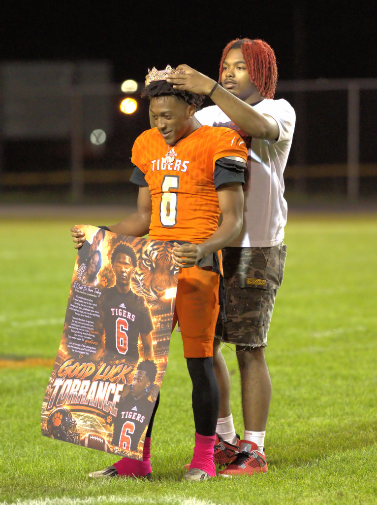 A Benton Harbor Tigers football player wearing jersey number 6 receives a crown during a homecoming ceremony while holding a personalized poster.