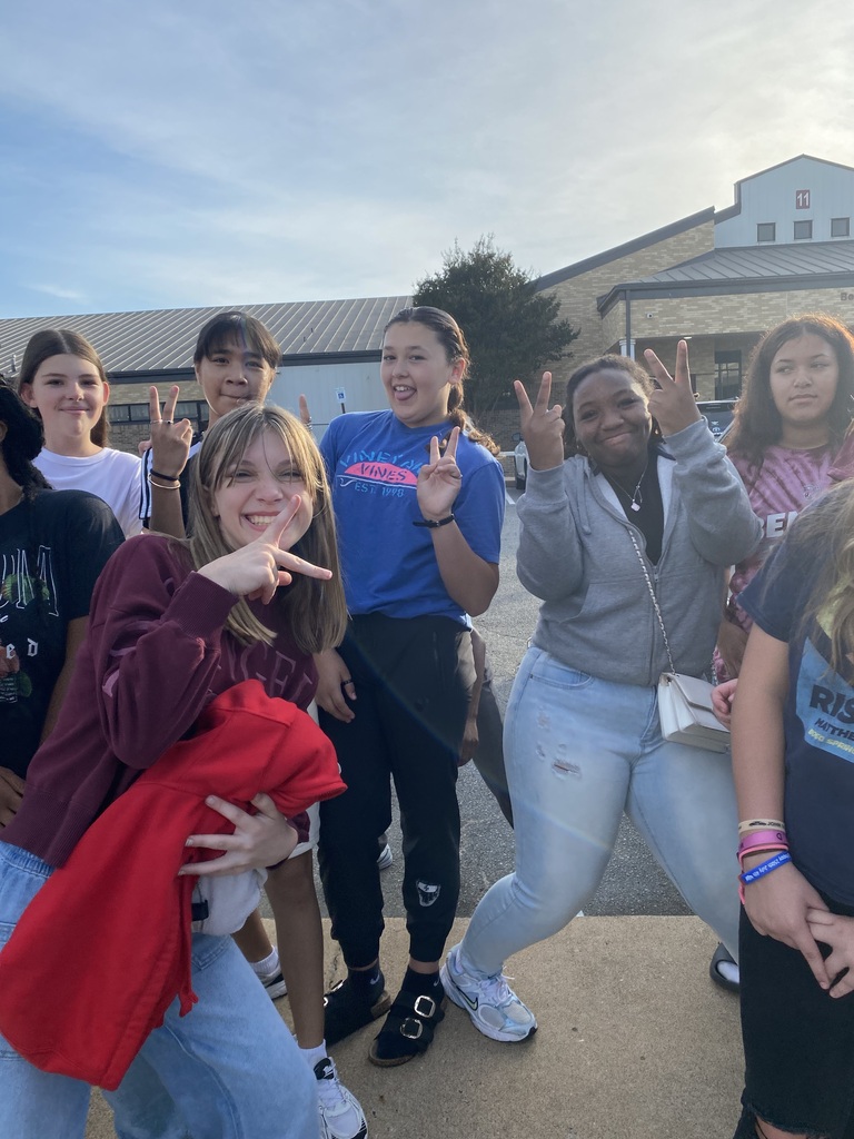A group of 6 students are in the frame displaying peace signs with their hands as they wait outside the middle school for the senior parade to start.