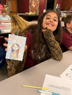 A student smiling at the camera as she displays artwork from class.
