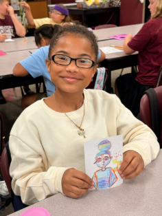 A student smiling at the camera as she displays artwork from class.