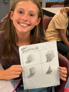 A student smiling at the camera as she displays artwork from class.