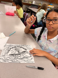 A student looks up at the camera with her artwork on the table in front of her. She is in the process of creating the art.
