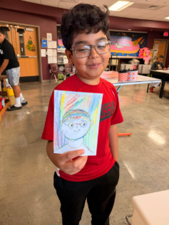 A student smiling at the camera as he displays artwork from class.