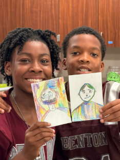 Two students smiling at the camera as they display their artwork from class.