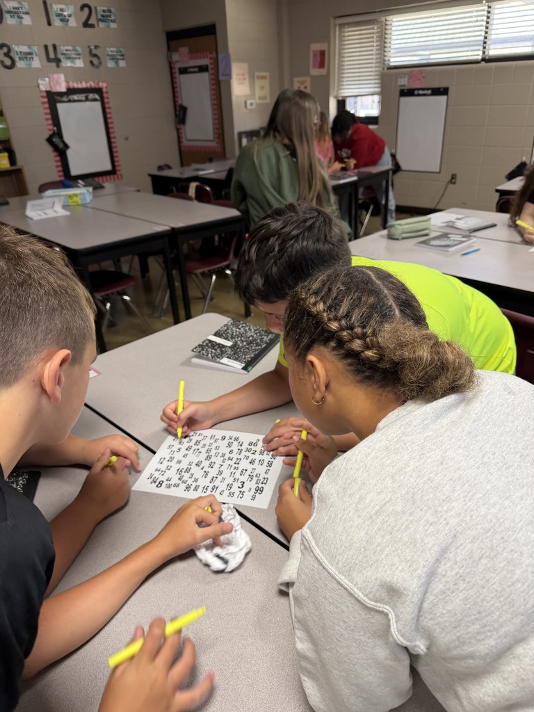 Three students huddle around a paper filled with different numbers as they work together to find numbers 1-100