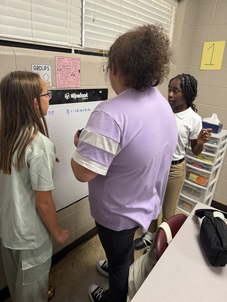 Three students work together to solve problems at chart paper on the wall of Mrs. Harris's Classroom.