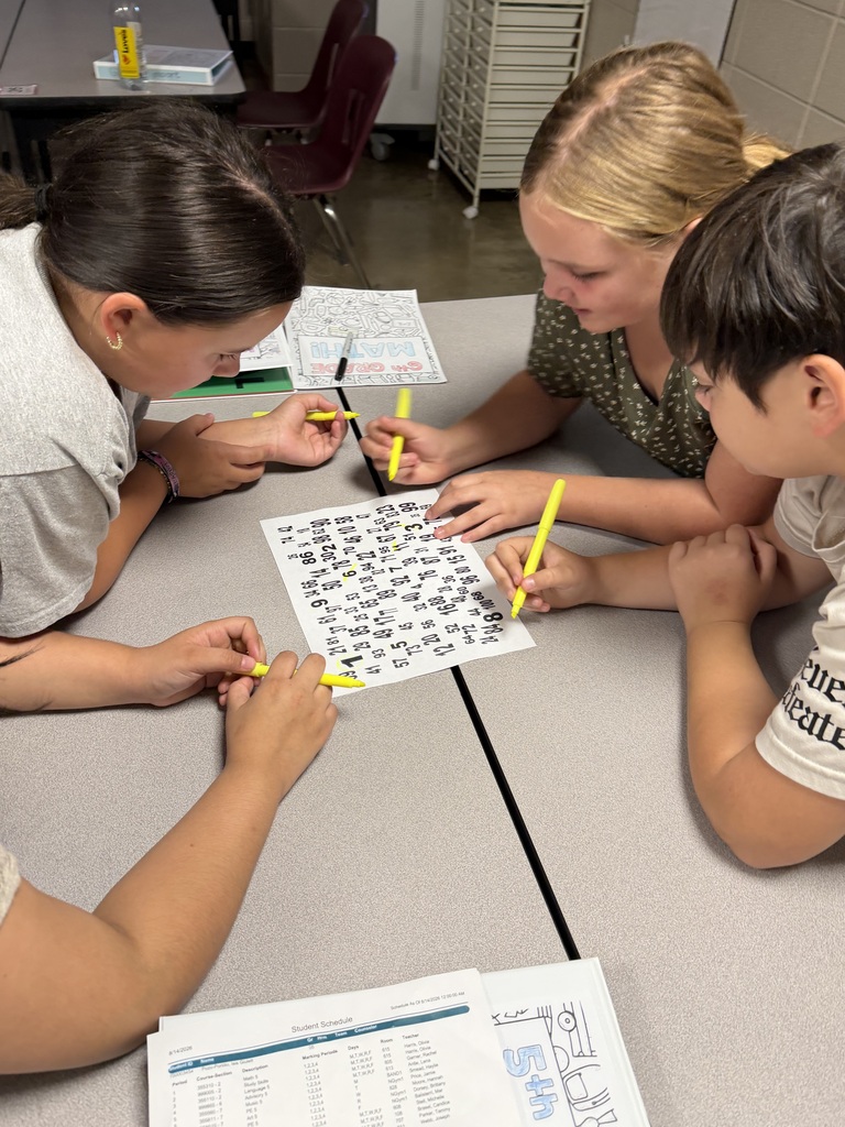 Three students huddle around a paper filled with different numbers as they work together to find numbers 1-100