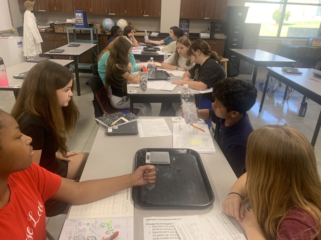 A group of students in Mr. Koch's class sit around their table with a plastic bottle and a digital scale as they watch a science experiment taking place.