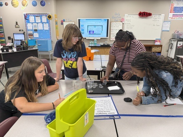 Students are standing around a table in Mrs. Eddleman's class watching a science experiment.