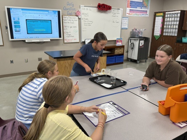 Students are standing around a table in Mrs. Eddleman's class watching a science experiment.