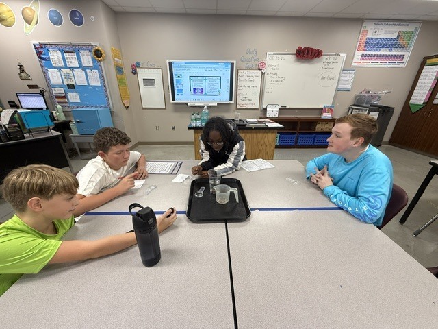 Students are standing around a table in Mrs. Eddleman's class watching a science experiment.
