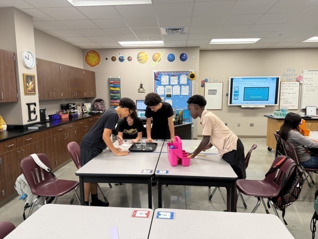 Students are standing around a table in Mrs. Eddleman's class watching a science experiment.