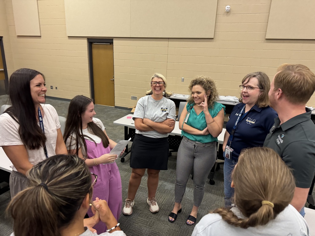 A group of new teachers and student leaders stand in a circle, smiling and laughing during a casual welcome activity in a classroom. The atmosphere is lively and warm, with participants holding papers and enjoying the interaction.