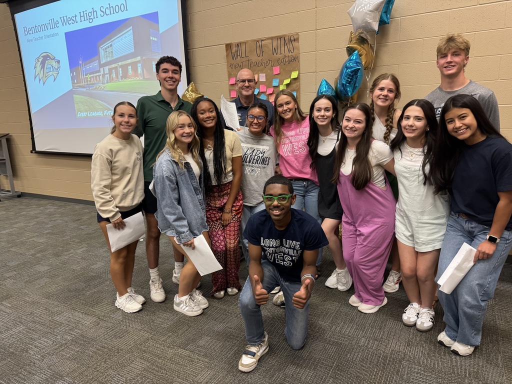 A group of new teachers and student leaders stand in a circle, smiling and laughing during a casual welcome activity in a classroom. The atmosphere is lively and warm, with participants holding papers and enjoying the interaction.