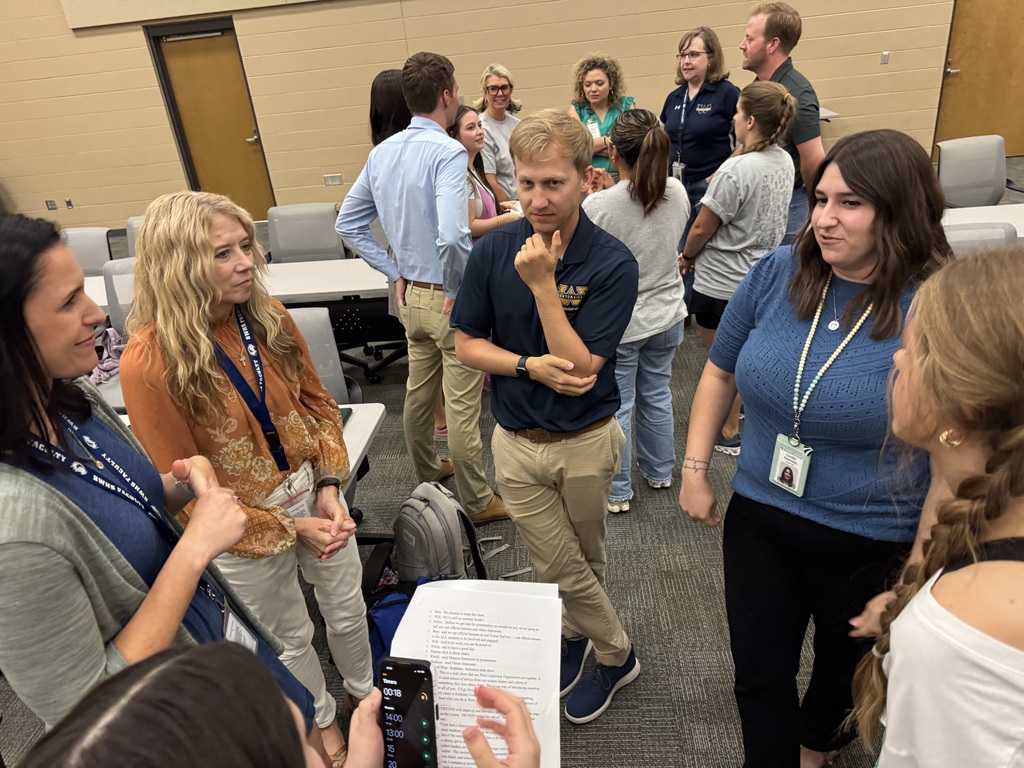 A group of new teachers and student leaders stand in a circle, smiling and laughing during a casual welcome activity in a classroom. The atmosphere is lively and warm, with participants holding papers and enjoying the interaction.
