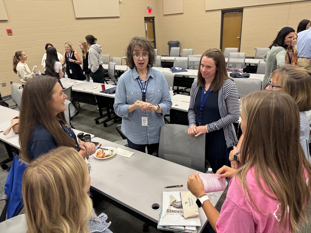 A group of new teachers and student leaders stand in a circle, smiling and laughing during a casual welcome activity in a classroom. The atmosphere is lively and warm, with participants holding papers and enjoying the interaction.