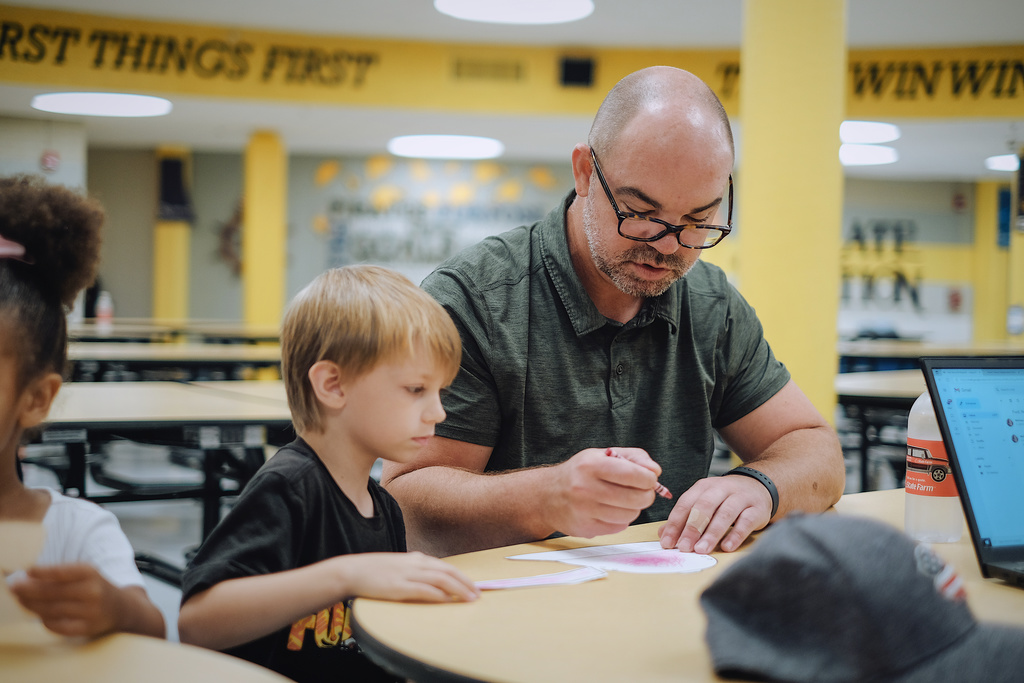 School Principal helps students craft at lunch table.