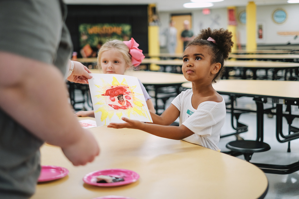 Student hands teacher a drawing while sitting at a lunch table