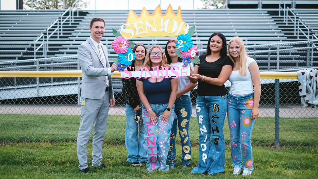 Superintendent poses with seniors on football field around a handmade frame that says "Senior Sunrise"