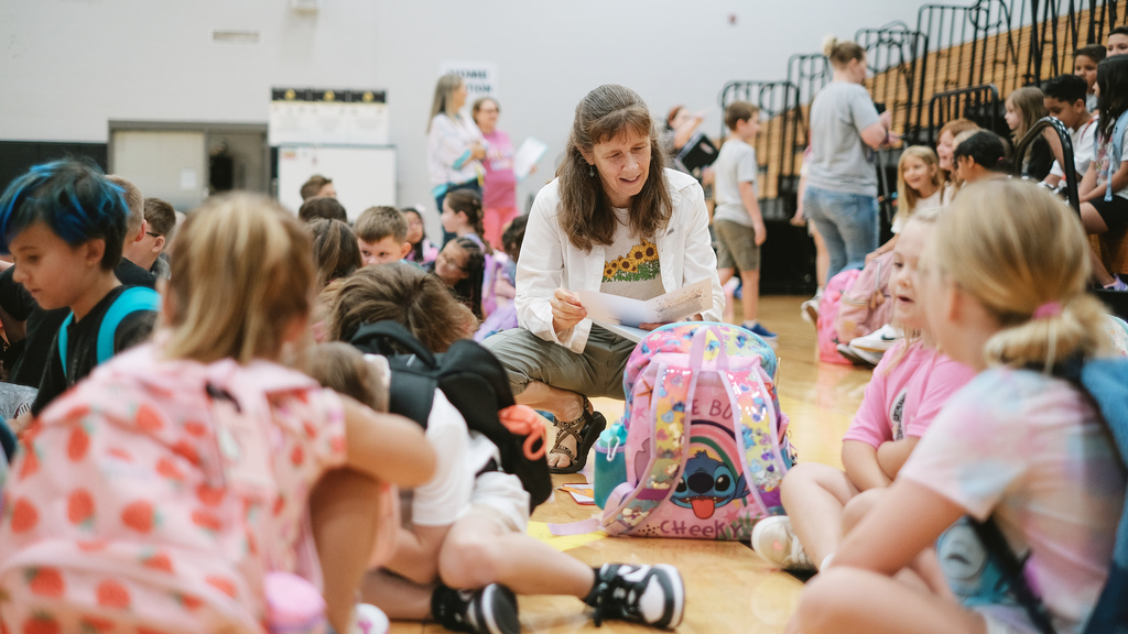 Elementary teacher kneels down next to a student on gym floor.
