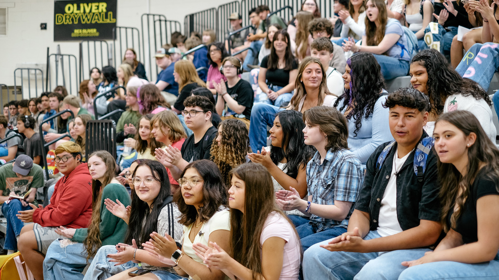 Students applaud for their teachers, seated on gym bleachers.