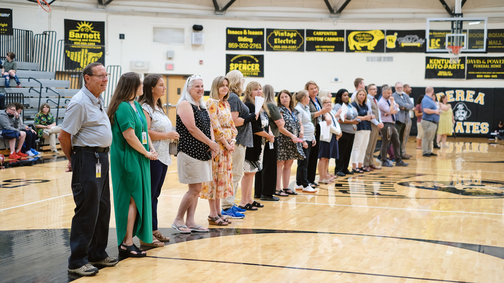 Teachers and staff stand in a line in front of students in school gym