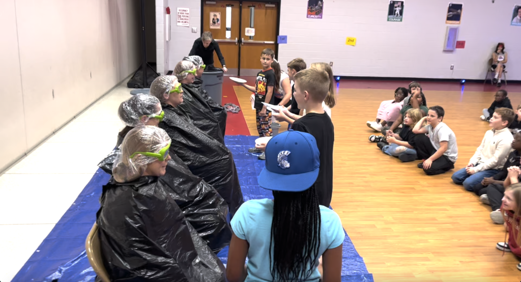 Image of 6 students pieing fourth grade teachers. Teachers sitting in chairs on blue tarp. Students holding white plates with whip cream! 