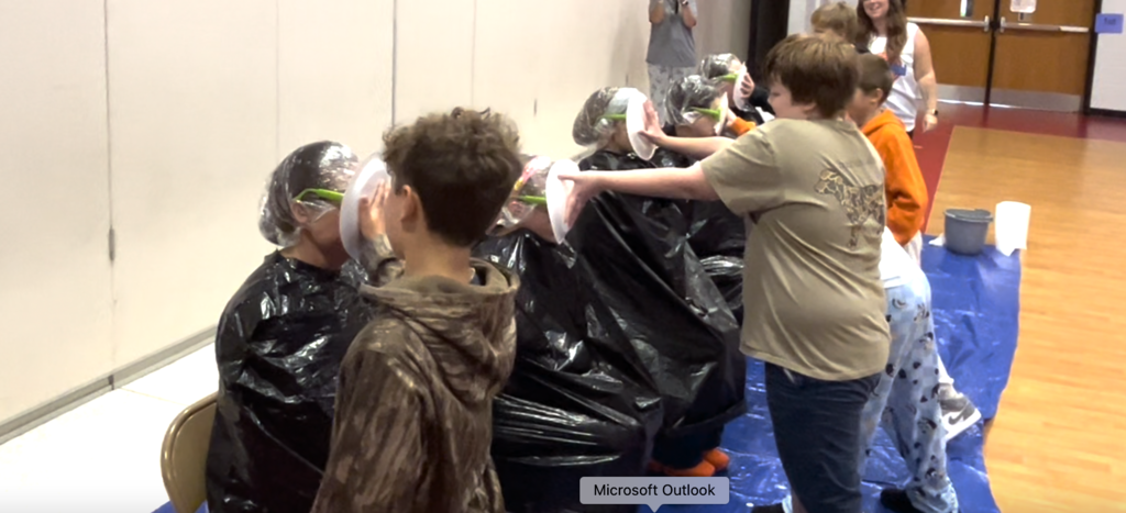 Image of 5 students pieing fifth grade teachers. Teachers sitting in chairs on blue tarp. Students holding white plates with whip cream! 