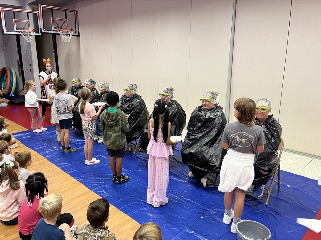 Image of 7 students pieing second grade teachers. Teachers sitting in chairs on blue tarp. Students holding white plates with whip cream! 