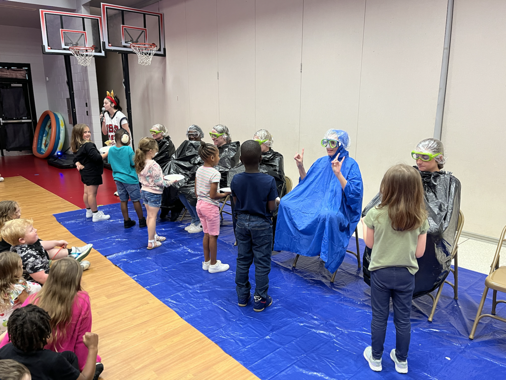 Image of 6 students pieing first grade teachers. Teachers sitting in chairs on blue tarp. Students holding white plates with whip cream! 
