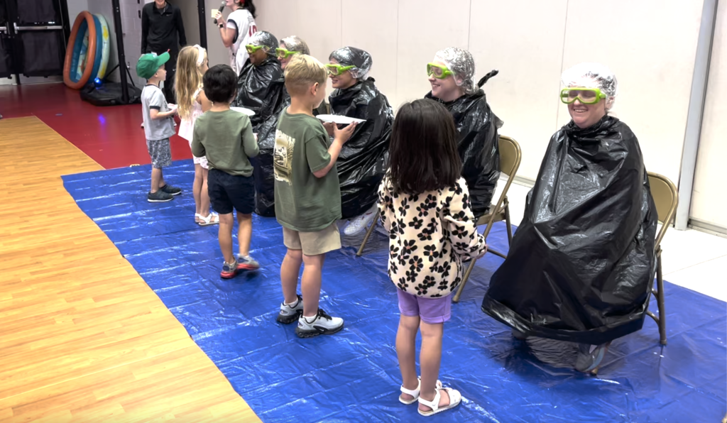 Image of 5 students pieing 5 K5 grade teachers. Teachers sitting in chairs on blue tarp. Students holding white plates with whip cream! 