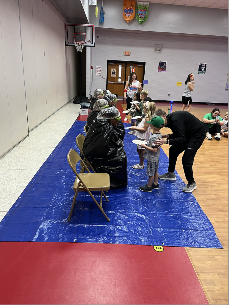 Image of 5 students pieing 5 K5 grade teachers. Teachers sitting in chairs on blue tarp. Students holding white plates with whip cream, image from the other side