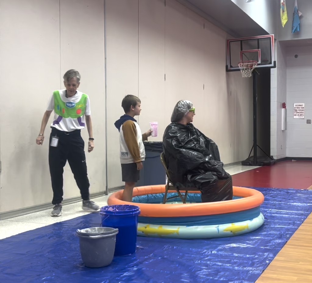 Image of a teacher sitting in a kiddie pool with a student standing behind her holding a cup of pink slime. 