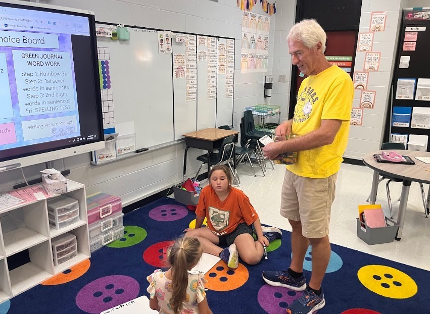 One of our volunteers working two students down on the carpet, spelling words on their whiteboards.