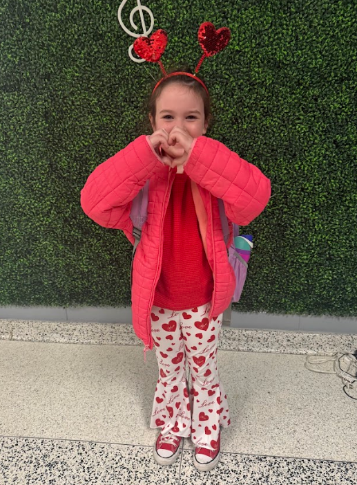 Little girl sports a red outfit with hearts on her pants and two hearts to match on her headband. She holds up the heart sign with her hands.