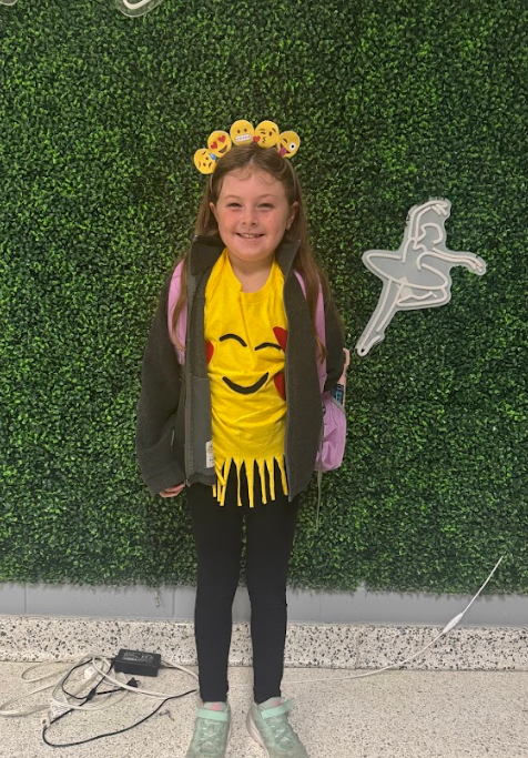Young girl dressed in a yellow shirt with a emoji smiley face surrounded with hearts and a headband that includes 5 different emoji smiley faces.