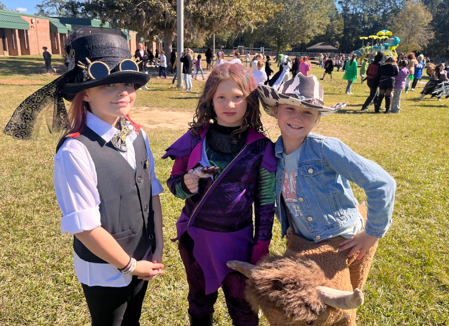 Three girls dressed in halloween costumes outdoors, posing together with big smiles. 