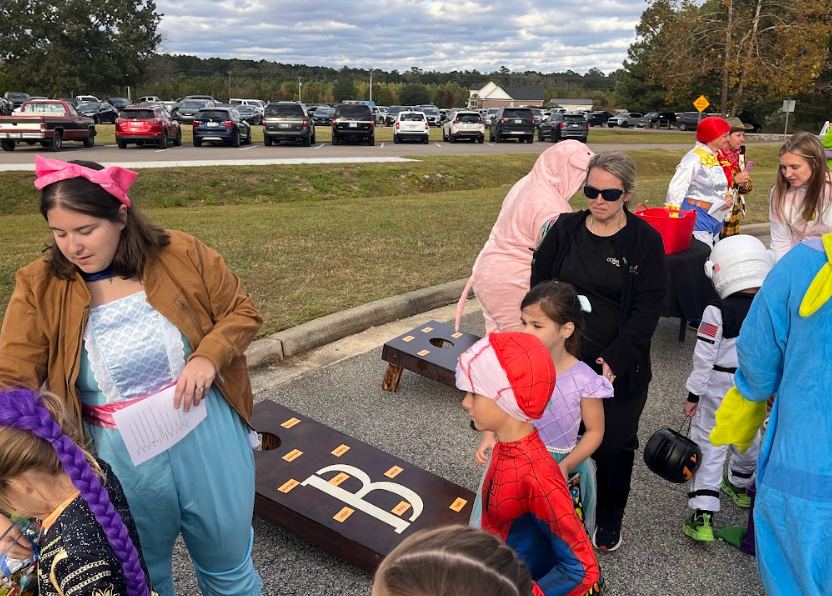Families stand in line for resources while others in the background are playing a game of cornhole where they read the word their bean bag lands on.