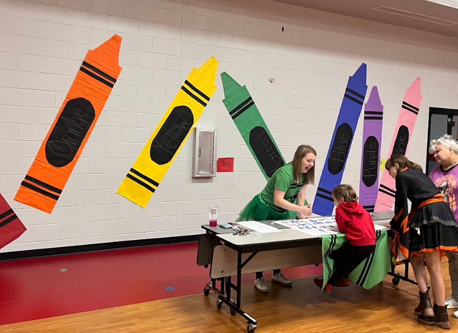 Teacher and family work together at a table to spell out words as they listen to the sounds of the letters. Crayons are drawn large int he background to go with the crayons theme for the kindergarten group.