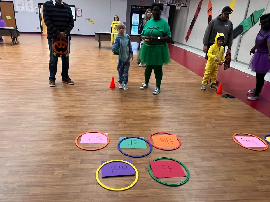 Student stands by teacher to throw bean bags in rings that have cvc and heart words from the ufli program. Where the bean bag lands, the student will read the word.