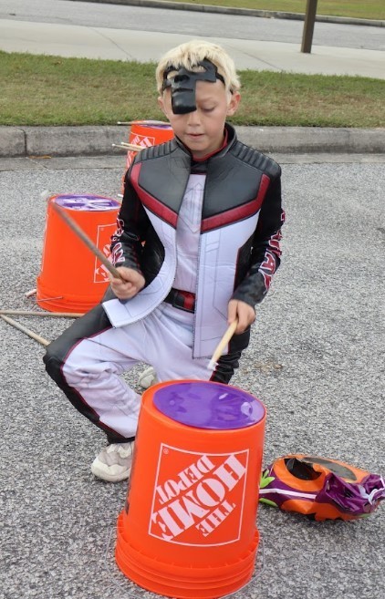 Student dressed in costume playing on a bucket with drumsticks.