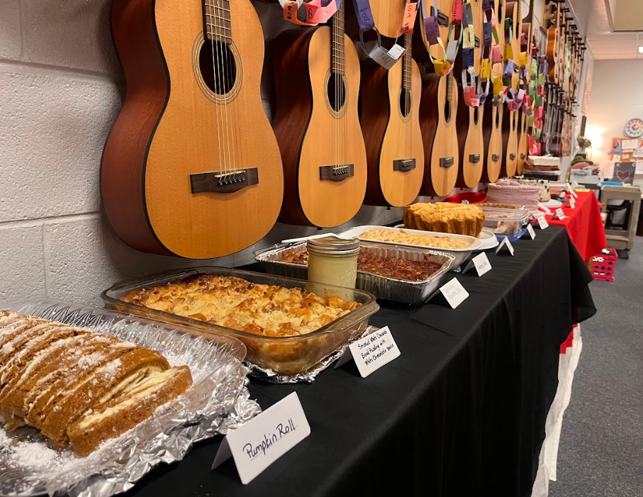This image includes a shelf of the baked goodies that were shared with HBE's faculty. There are a number of desserts with tags in front of them listing what they are. Guitars hang above the desserts because it was hosted in the music room.