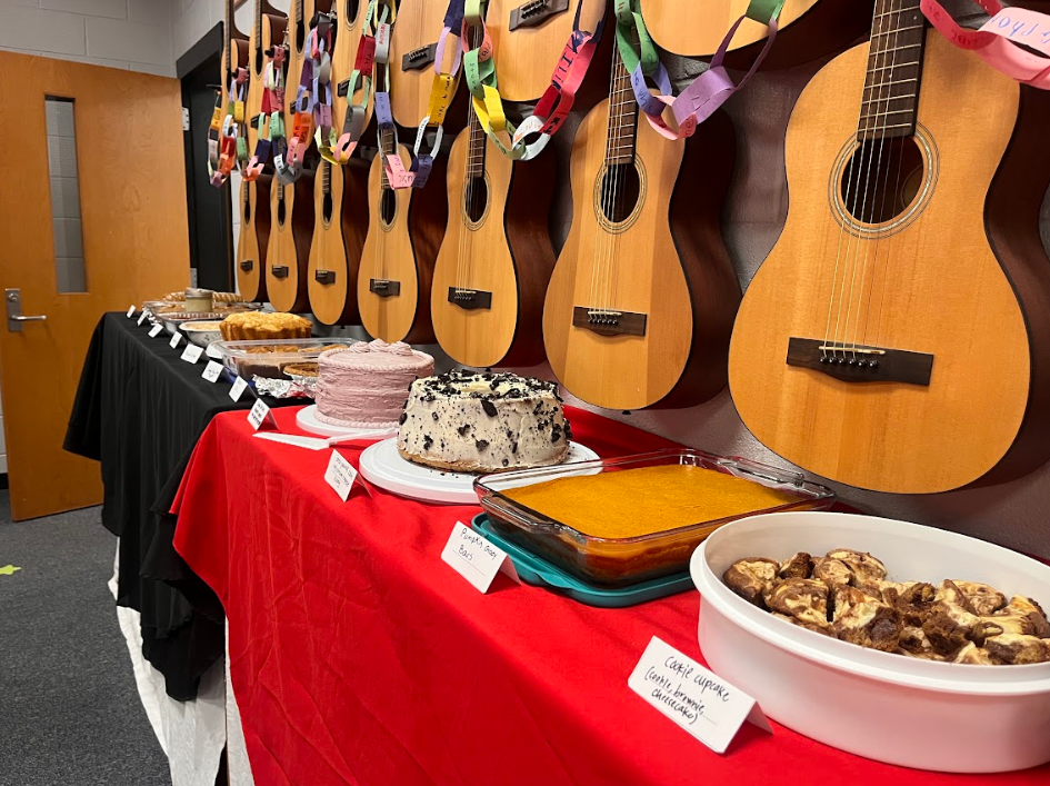 This image includes a shelf of the baked goodies that were shared with HBE's faculty. There are a number of desserts with tags in front of them listing what they are. Guitars hang above the desserts because it was hosted in the music room.
