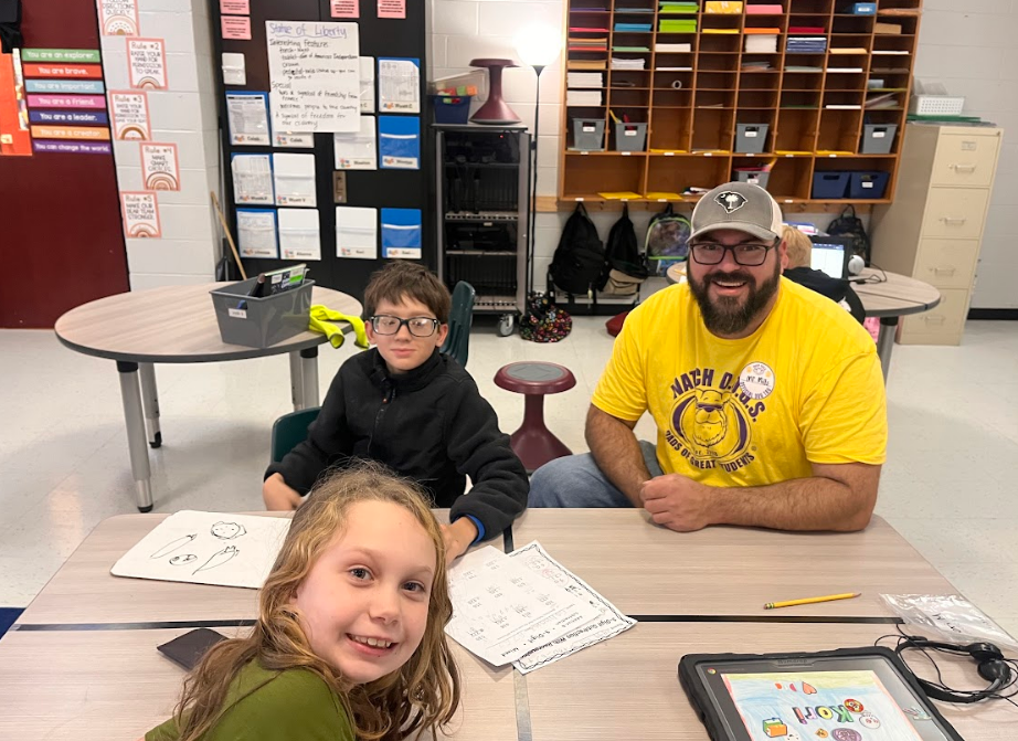 A young boy, young girl, and male volunteer in a Watch DOGS t-shirt pose for a picture at a table. Student work is in the center of the table showing they were doing some work prior to saying cheese.